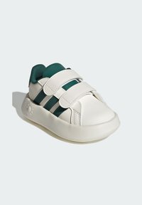 Zapatilla blanca con acentos verdes, parte superior sintética, correas de velcro, punta redonda y suela de goma texturizada. Presenta tres rayas horizontales.