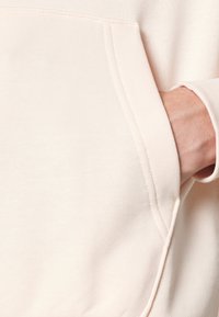 Ljust rosa sweatshirt med en kenguruficka, ribbad mudd och en mjuk bomullstextur. Synlig söm runt fickan.