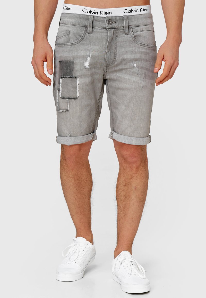 Sivi traper šorts s razderanim rubom, s detaljima zakrpa i istrošenim naglascima. Calvins Klein logo na pojasu, ležeran kroj. Bijele tenisice.