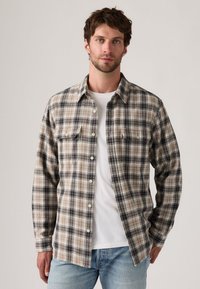 Flanellskjorte i beige og svart rutemønster, med lange ermer, to brystlommer og hvite knapper, lagd over en hvit t-skjorte.