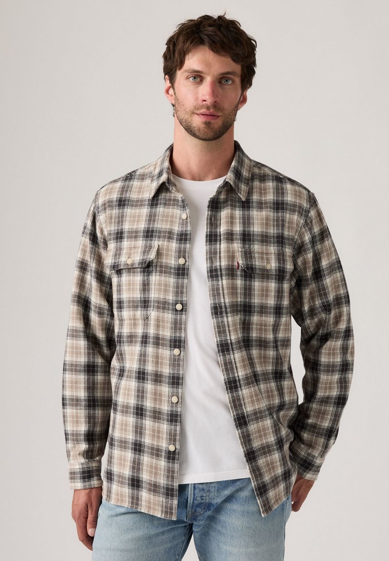Flannel shirt in beige and black check pattern, featuring long sleeves, two chest pockets, and white buttons, layered over a white t-shirt.