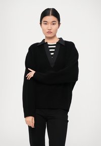 Schwarzer, oversized Strickpullover mit tiefem V-Ausschnitt und kontrastierendem schwarzen Kragen. Über einem schwarz-weiß gestreiften Oberteil getragen.