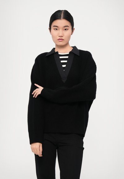 Black knitted oversized sweater with a deep V-neck and contrasting black collar. Worn over a black and white striped top.
