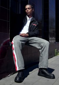 Chaqueta negra con cremallera y logo rojo, camiseta polo blanca, jeans grises con franjas laterales rojas y zapatillas negras. Sentado contra azulejos oscuros.