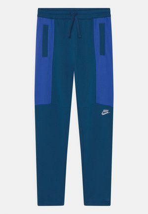 Marineblå og blå træningsbukser med snoretræk i taljen, sidelommer og et lille sølvfarvet Nike-logo på det nederste ben. Blødt, glat stof.