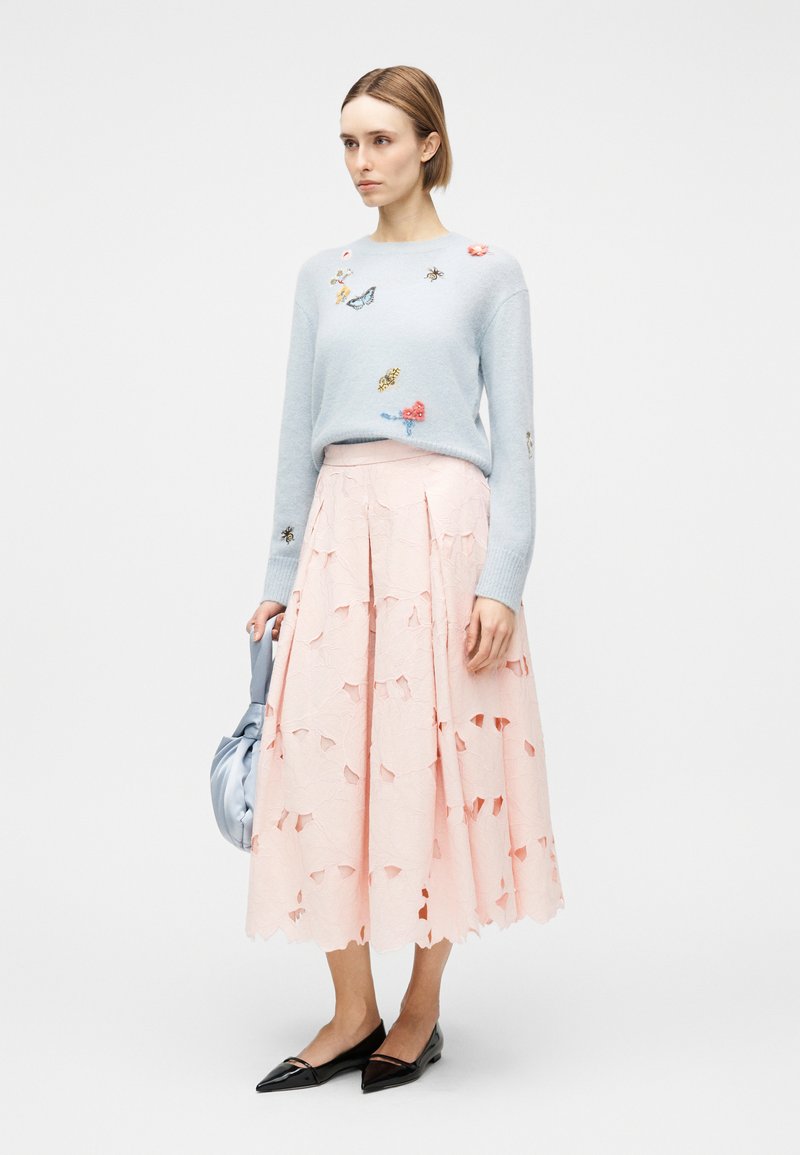 Donna che indossa un maglione azzurro chiaro con ricami di farfalle e insetti, gonna midi rosa con motivi traforati, scarpe basse nere, che tiene una borsa azzurro chiaro.