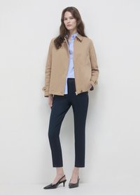 Veste extérieure beige avec col, portée sur une chemise bleu clair. Pantalon bleu marine coupe slim et escarpins noirs à talons aiguilles complètent la tenue.