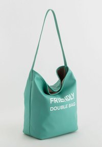 Turkos konstläder axelväska med en stor öppning, "FRIENDLY DOUBLE BAG" tryckt i vitt och en mjuk, strukturerad finish.