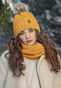 Yellow knitted beanie with a pom-pom on top, featuring a textured pattern. Paired with a matching yellow knitted scarf and a cream jacket.