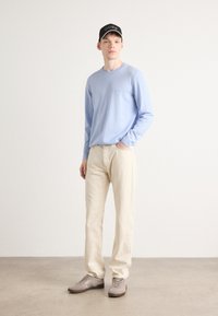 Camisa de manga longa azul-claro, calças bege e sapatilhas cinzentas. Apresenta um gola redonda e um bordado discreto de logotipo na camisa.