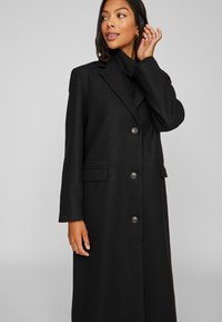 Pikend must jope, mis on valmistatud tekstuuriga kangast, millel on nurgelised kraed, viis nööpi ja küljetaskud. Klasikaline siluett ja kohandatud disain.