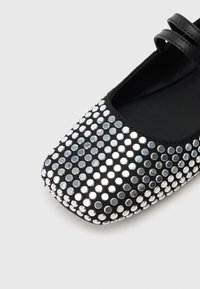 Musta litteä kenkä pyöreällä kärjellä, joka on koristeltu hopeisilla strasseilla pistekuviossa ja jossa on musta hihna koko ylle.