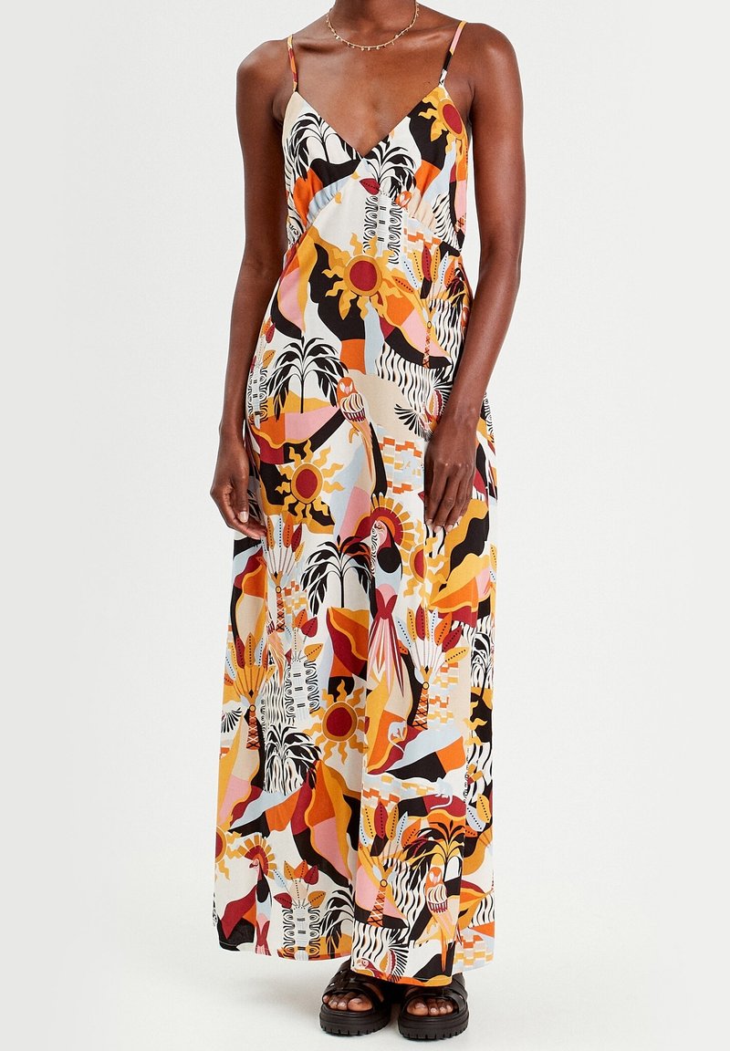 Femme portant une robe maxi sans manches à col en V avec des motifs tropicaux abstraits en orange, noir, jaune, rose et blanc, associée à des sandales noires.