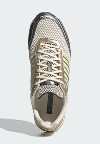 Αθλητικά παπούτσια με επάνω μέρος από διχτυωτό υλικό σε μπεζ και γκρι, μαύρες λεπτομέρειες, κίτρινες ρίγες και καουτσούκ σόλα· διαθέτουν κλείσιμο με κορδόνια και επενδεδυμένο πέλμα.