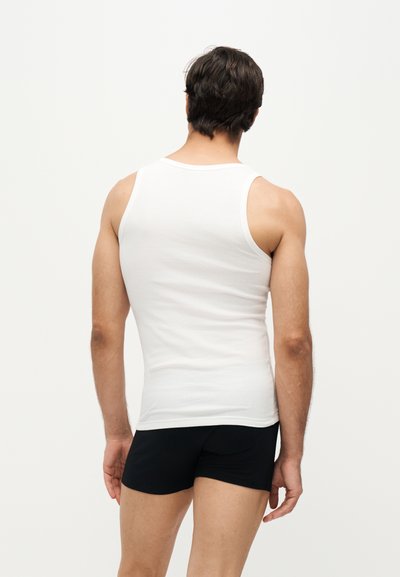 Homme portant un débardeur blanc et un short noir, debout, dos à la caméra, sur un fond blanc uni.
