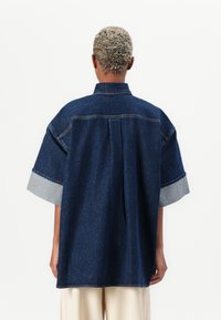 Blue denim shirt with short sleeves, featuring a collared neckline and light blue cuffs. Relaxed fit with a back pleat and minimal stitching.