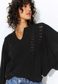 Haut noir avec un décolleté en V, tissu tricoté texturé, détails en dentelle sur les côtés, et manches fluides, associé à un short en denim bleu clair.