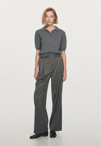 Donna in piedi che indossa un polo a maniche corte grigio in maglia e pantaloni a gamba larga a righe con vita alta, accompagnati da scarpe nere.