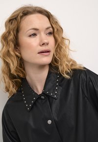 Melnas ādas jaka ar lielu apkakli, kas rotāta ar sudraba kniedēm, ar spiedpogām un gludu tekstūru. Modelim ir viļņaini, plecu garuma mati.