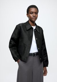 Μαύρο σακάκι με μοτίβο, γιακά, κλείσιμο με κουμπιά και ελαστικές μανσέτες, σε συνδυασμό με λευκό πουκάμισο και γκρι παντελόνι.