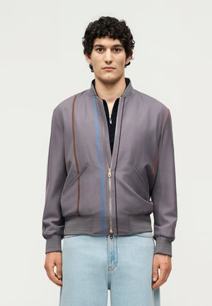 Giovane uomo con capelli ricci indossa una giacca grigia con zip e strisce verticali marroni e blu, abbinata a jeans azzurri, in piedi in modo neutro.