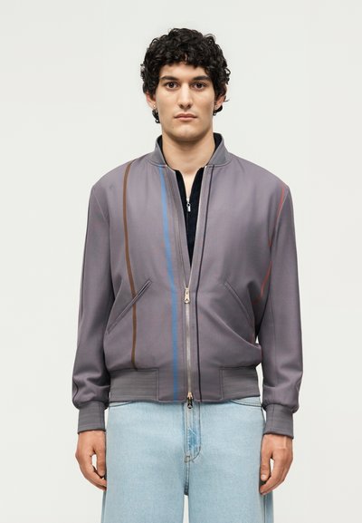 Jeune homme aux cheveux bouclés porte une veste grise zippée avec des rayures verticales marron et bleues, associée à un jean bleu clair, debout dans une posture neutre.