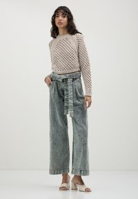 Donna che indossa un maglione beige all'uncinetto, jeans a vita alta a gamba larga con effetto acid-wash e cintura, e sandali con tacco a blocco open-toe color crema, in piedi contro un muro semplice.