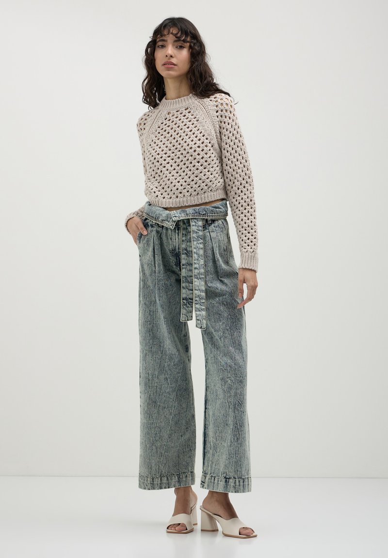 Donna che indossa un maglione beige all'uncinetto, jeans a vita alta a gamba larga con effetto acid-wash e cintura, e sandali con tacco a blocco open-toe color crema, in piedi contro un muro semplice.