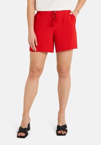 Shorts rouges texturés avec une taille à cordon de serrage et des poches latérales, associés à des sandales noires à talons carrés avec des brides croisées.