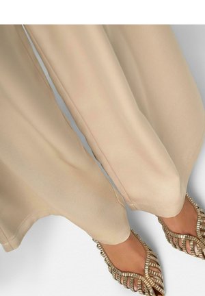 Voeten met beige geweven open teen platte schoenen gecombineerd met lichtbeige brede broekspijpen tegen een witte achtergrond.