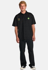 Black short-sleeve polo shirt with a geometric pattern, featuring green, red, and yellow logo accents on the chest. Pair of black pants.