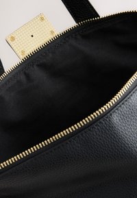 Sac en cuir noir avec fermeture éclair dorée ouverte montrant l'intérieur en tissu noir vide et un fermoir doré avec des vis.