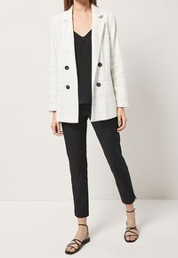 Vrouw in een wit dubbelzijdig geruit blazer, zwarte mouwloze top, zwarte strakke broek en zwarte sandalen met bandjes die tegen een effen muur staat.