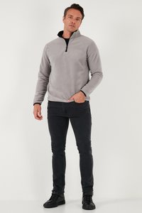 Vyras stovintis priešais baltą foną, dėvintis užtrauktą pilką flisinę striukę, juodus siaurus džinsus ir juodus aukštus sportinius batelius, viena ranka kišenėje.