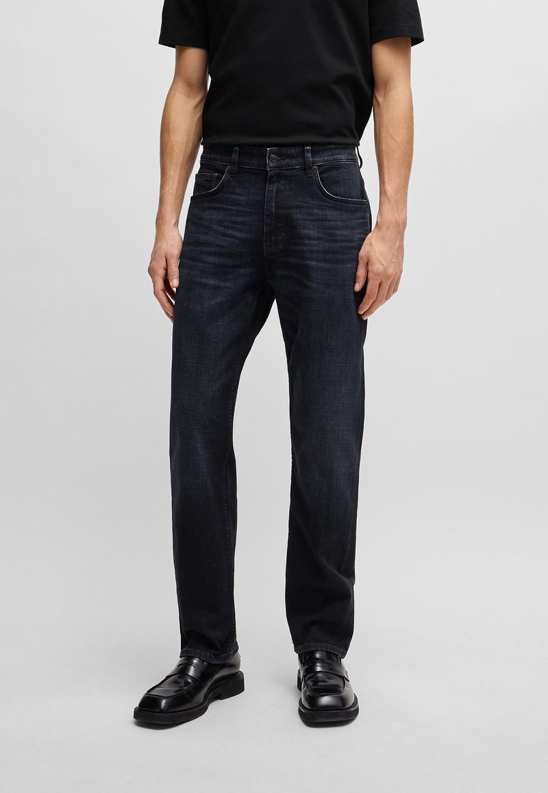 Jeans in denim blu scuro con taglio dritto, cinque tasche, sfumature leggere e dettagli di cucitura chiari. Indossati con scarpe nere e una camicia.