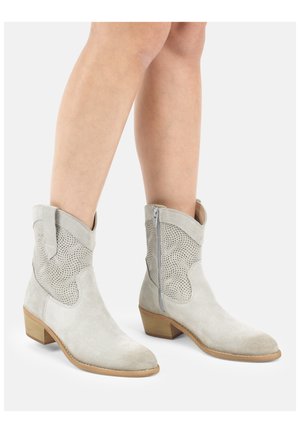 Lichtgrijze suède enkellaarsjes met lage houten hakken, ritssluitingen aan de zijkant en geperforeerde panelen met spiraalpatroon, gedragen op blote benen.