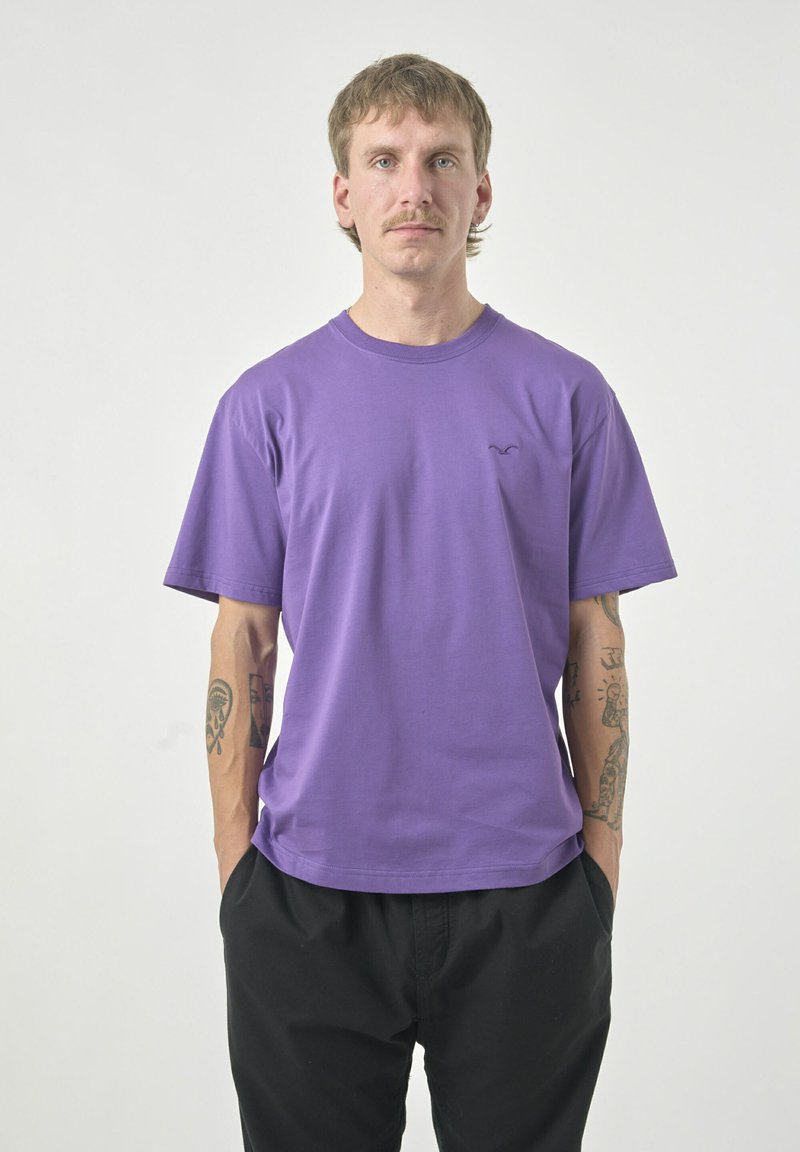Purple cotton crew-neck t-shirt with short sleeves and a small logo on the left chest, paired with black pants. Smooth texture.
