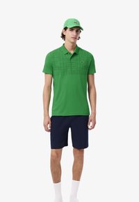 Uomo che indossa una polo a trama verde, un cappellino verde abbinato, pantaloni navy e calze bianche mentre è in piedi su uno sfondo bianco uniforme.
