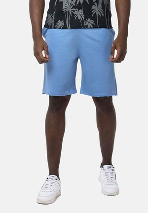 Uomo con pantaloncini casual azzurri chiari, t-shirt nera con stampa di palme grigia e sneakers bianche, in piedi con le mani rilassate.