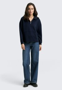Pull bleu marine avec un col zippé demi-long, manches longues et texture côtelée, associé à un jean en denim à jambes larges et des bottes noires.