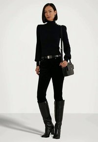 Γυναίκα που στέκεται φορώντας μαύρο πουλόβερ με ψηλό λαιμό, μαύρο παντελόνι, μαύρες μπότες μέχρι το γόνατο και κρατώντας μια μικρή μαύρη τσάντα ώμου.