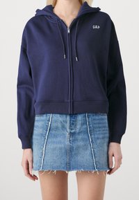 Sudadera de cremallera en color navy, fabricada en tejido suave, con capucha, que presenta un sutil logo de "GAP" y cordones ajustables, combinada con una falda mini de mezclilla deshilachada.