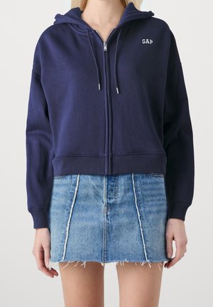 Sweat à zip navy en tissu doux avec capuche, arborant un logo "GAP" discret et des cordons de serrage réglables, associé à une mini-jupe en denim effilochée.