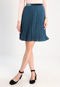Una gonna plissettata color turchese realizzata in un tessuto leggero, con un design testurizzato e una vita liscia e scura. Abbinata a scarpe con cinturino alla caviglia nere.