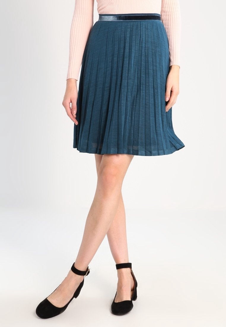 Una gonna plissettata color turchese realizzata in un tessuto leggero, con un design testurizzato e una vita liscia e scura. Abbinata a scarpe con cinturino alla caviglia nere.
