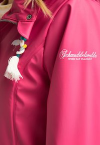 Giacca impermeabile rosa con zip, dotata di un cordino decorativo con pon pon e un logo stampato sulla manica. Texture liscia.