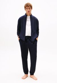 Jaunas vyras basas, apsirengęs tamsiai mėlyna striuke su užtrauktuku, derančiomis sportinėmis kelnėmis ir balta Tommy Hilfiger marškinėliais, stovintis prieš baltą foną.