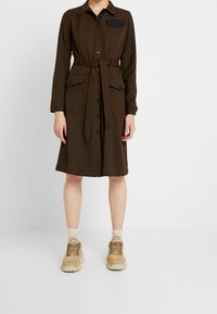 Ruskea nappikauluksellinen mekko, jossa on pitkät hihat, vyötäröllä vyö, kaksi etutaskua ja tumma sininen rintatasku, yhdistettynä vaaleisiin lenkkareihin.