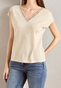 Beige V-hals top med korte ærmer, med blonder langs udskæringen. Stoffet ser blødt ud med en let glans. Kædet med blå jeans.
