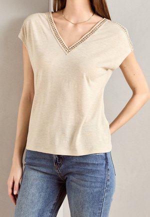 Beige V-Ausschnitt-Bluse mit kurzen Ärmeln, die eine Spitzenborte am Ausschnitt aufweist. Der Stoff wirkt weich mit einem leichten Glanz. Kombination mit blauen Jeans.